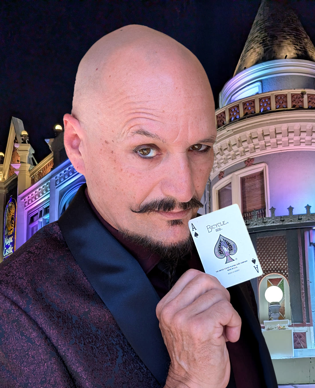 Ace Miles today, bald with a mustache and goatee, holding an ace of spades playing card in front of the lit Magic Castle turret in Hollywood at night.