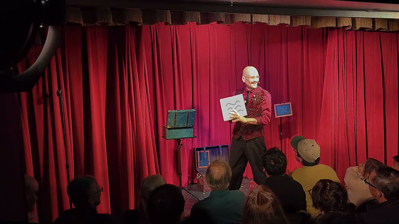 Ace Miles on stage holding a white prediction box in front of him, red curtain behind, audience watching from the front rows.