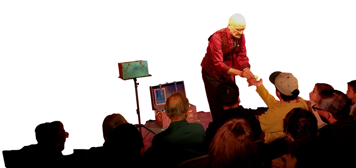 Ace Miles on a theater stage in a patterned red vest, handing a small object down to a boy in the front row, red curtain and prop trunks behind him, audience watching.