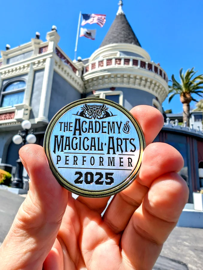 Ace Miles holding an Academy of Magical Arts performer coin, 2025, in front of the Magic Castle turret in Hollywood.